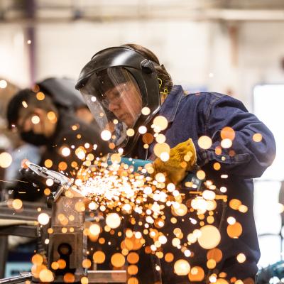 A student wearing protective gear, including a face shield and gloves, is using a welding tool, with bright sparks flying in the foreground. The student is focused on their work in a workshop environment.